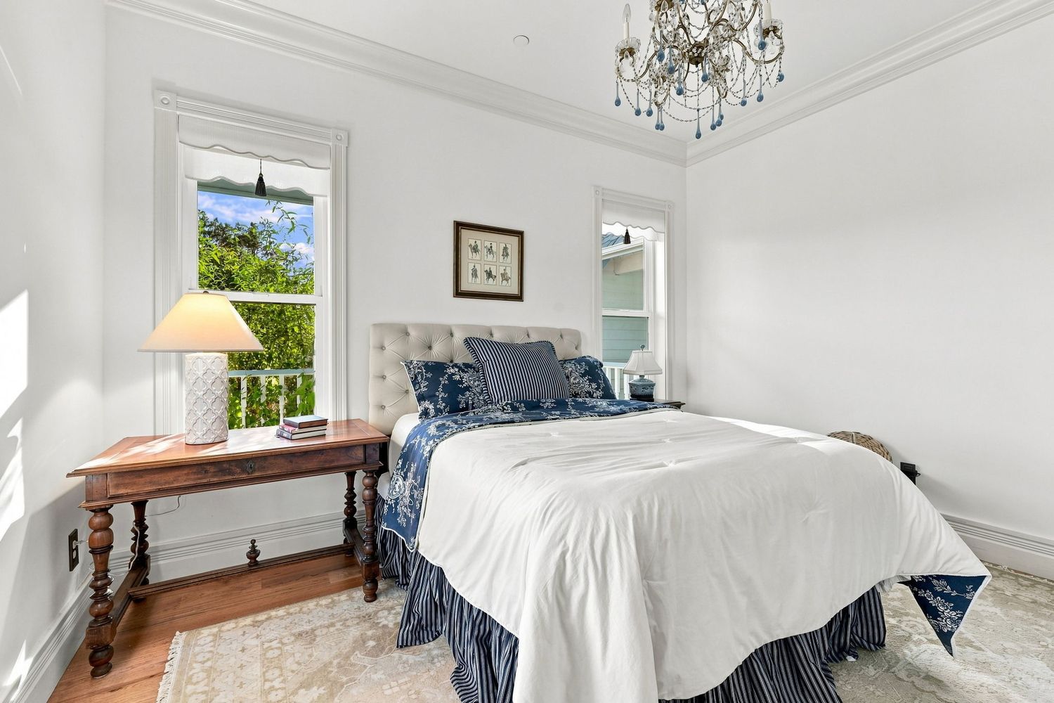Elegant white bedroom with blue accent bedding, crystal chandelier, hardwood floors, and a window overlooking lush greenery.