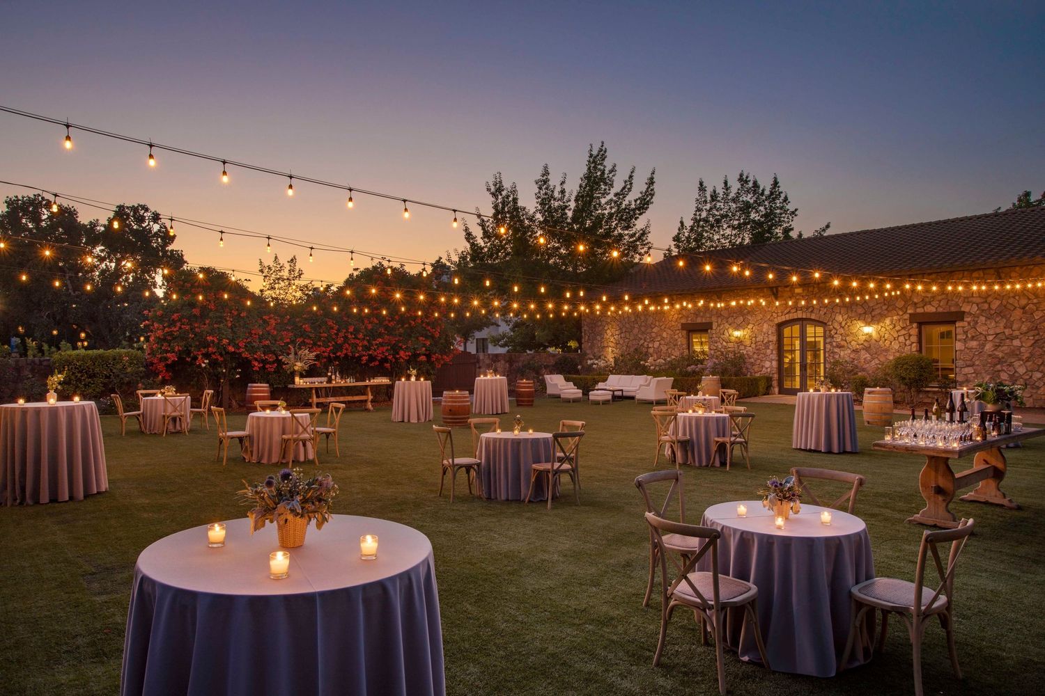String lights illuminate round tables with white linens at an elegant outdoor evening reception venue at sunset.