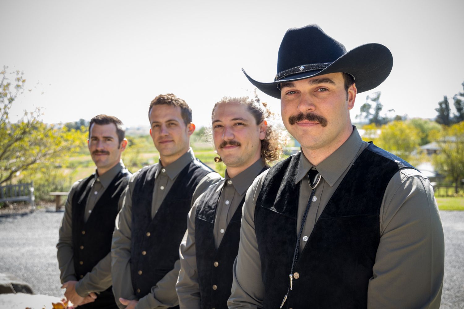 Four people wearing black vests and cowboy attire pose together outdoors against a sunny natural landscape.