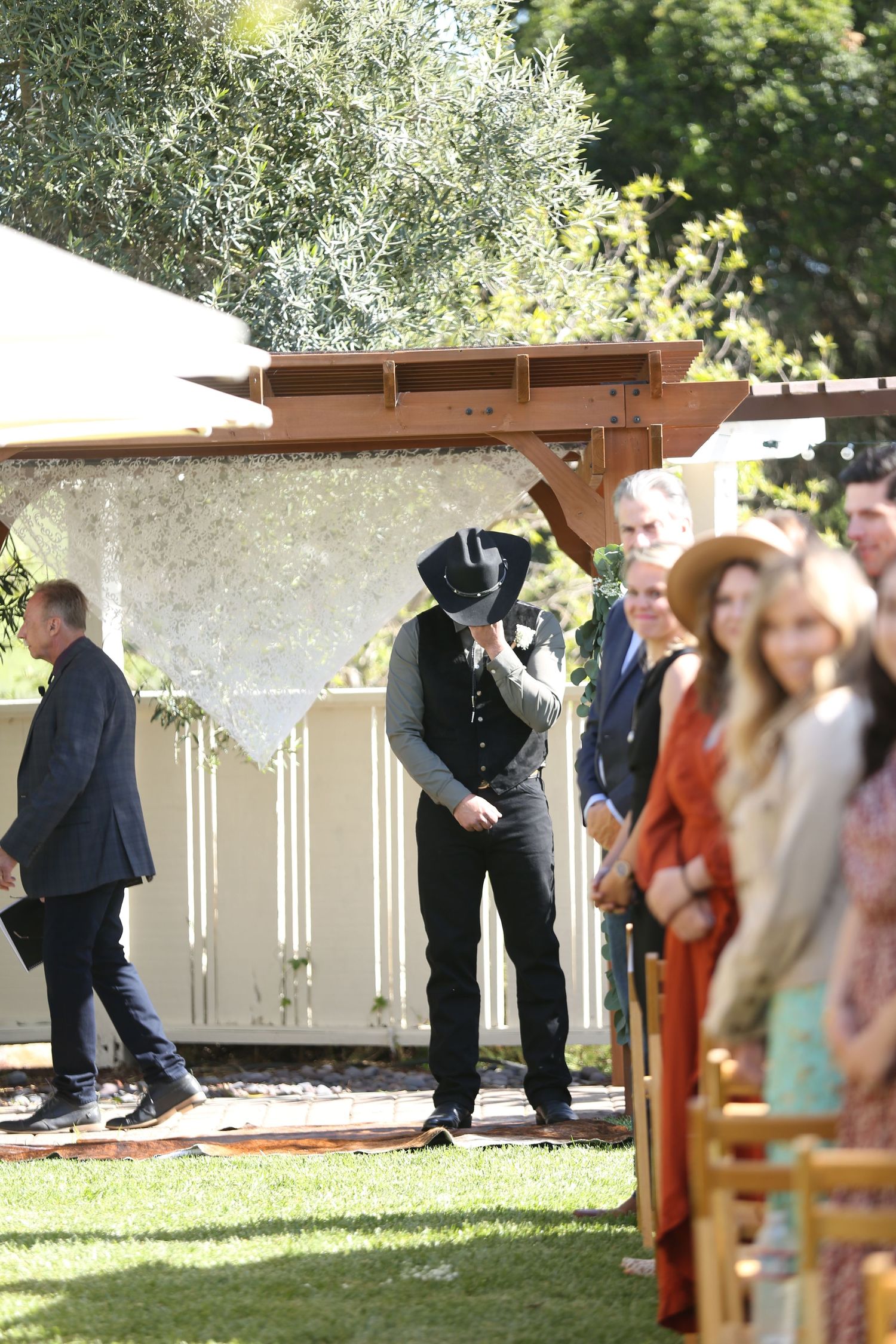 Wedding ceremony guests gather outdoors under a wooden pergola decorated with soft greenery and white floral arrangements.