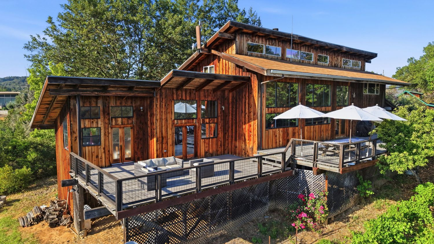 Rustic cedar wood home with large deck, outdoor dining area, and lush green surroundings on a sunny day.