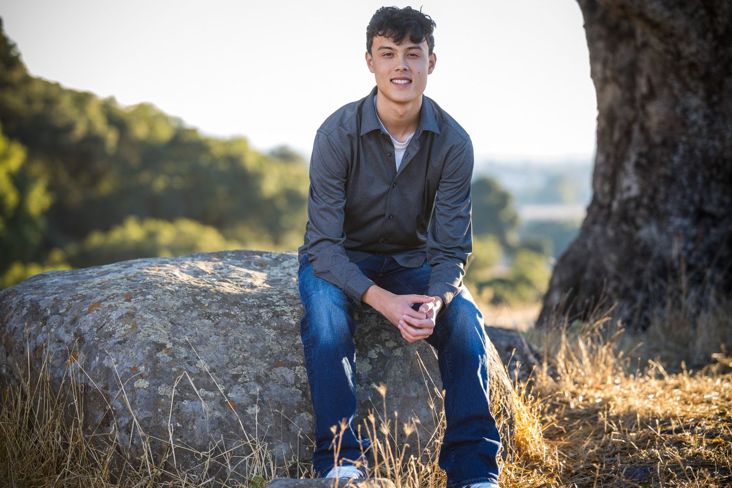 Senior Photos - Outdoor portrait of a person sitting on a rock next to a large tree with scenic background.