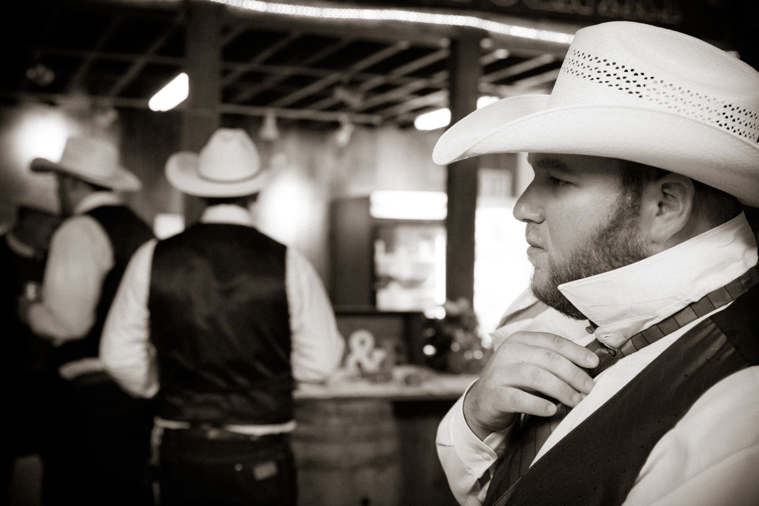 A black and white photo of a cowboy wearing a white hat and vest standing in a dimly lit western bar.