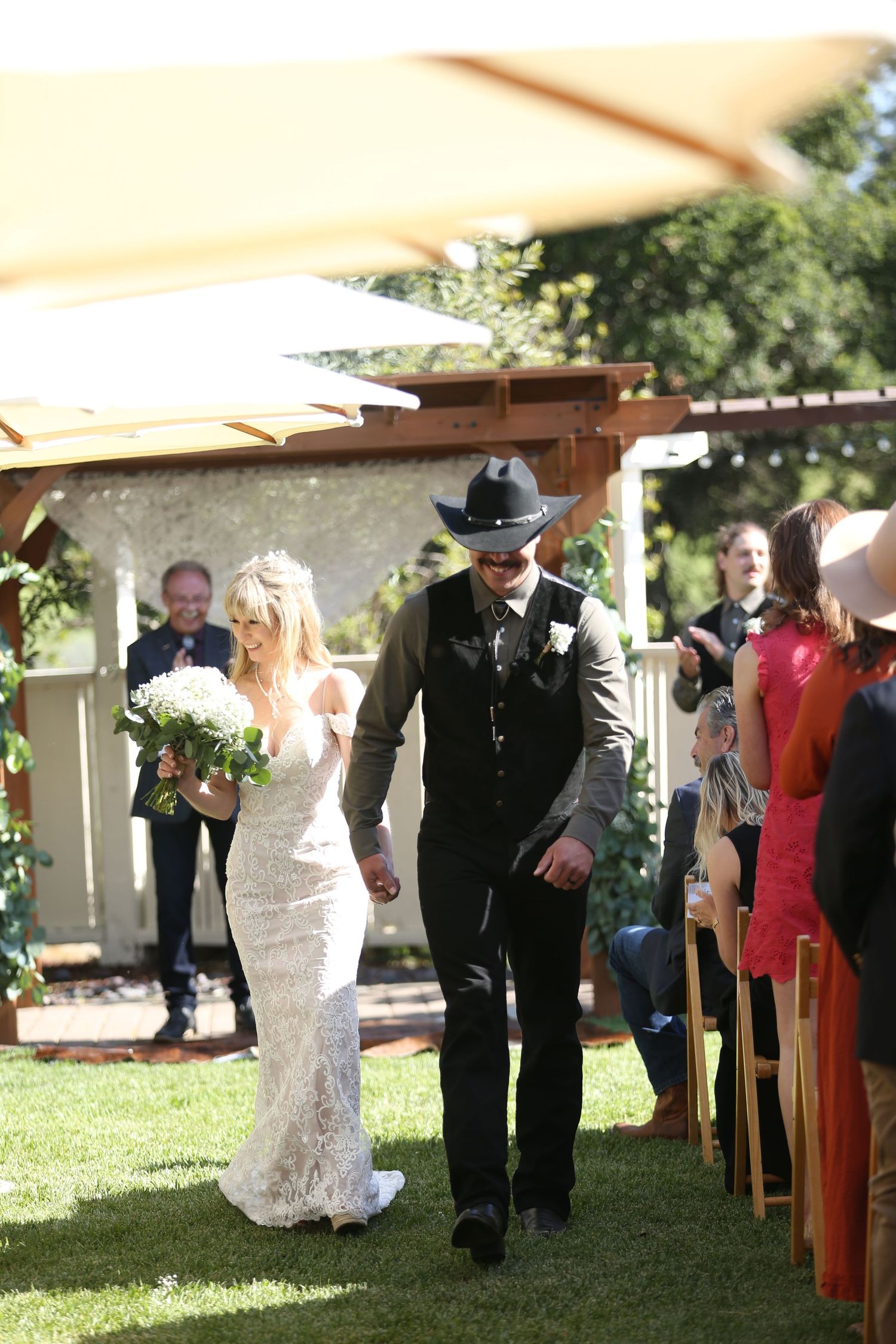 A wedding couple walks down an outdoor aisle under a wooden pergola at a garden ceremony.