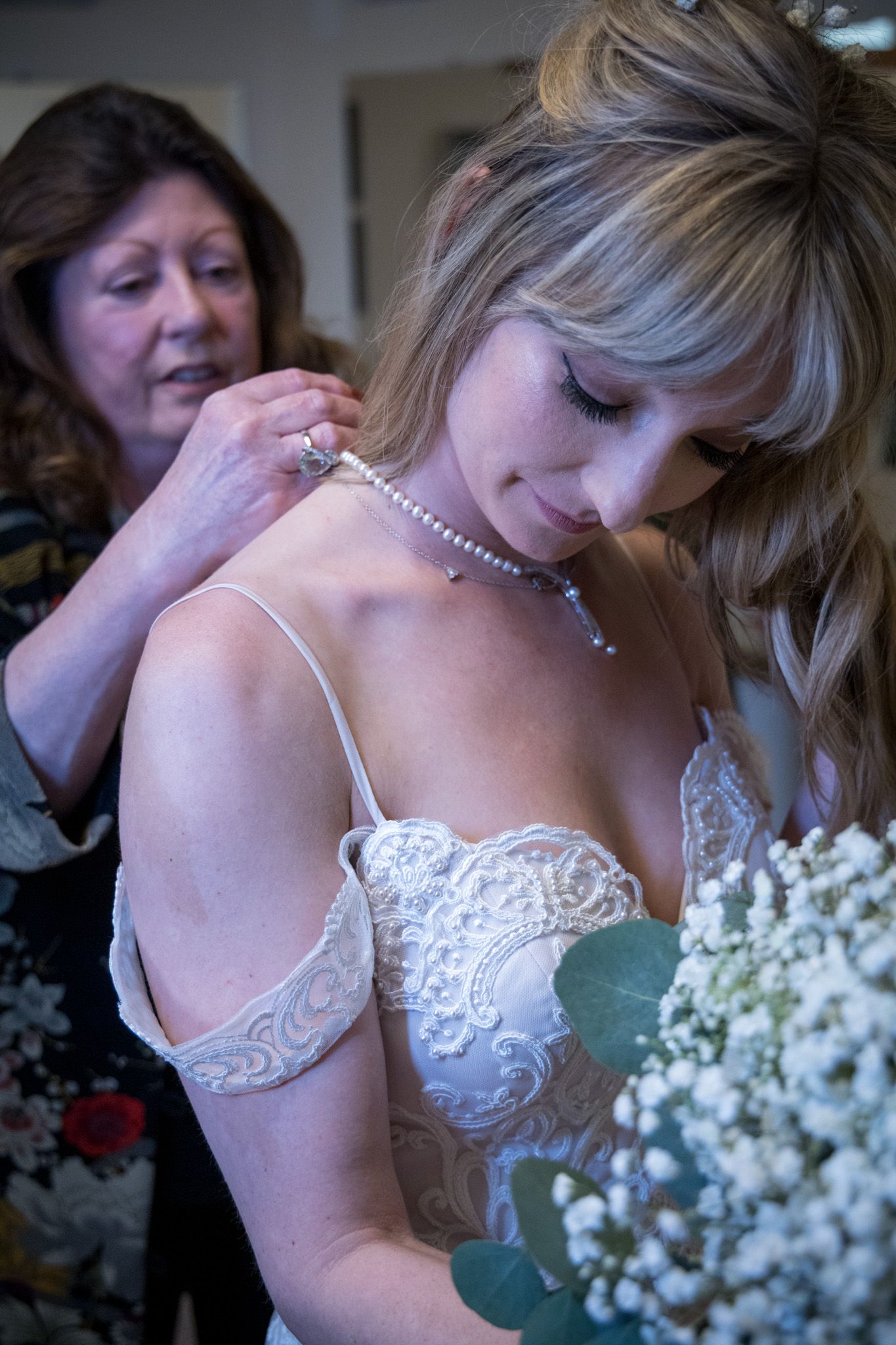 Pearl necklace being fastened onto a bride wearing a delicate lace wedding dress with baby's breath flowers.