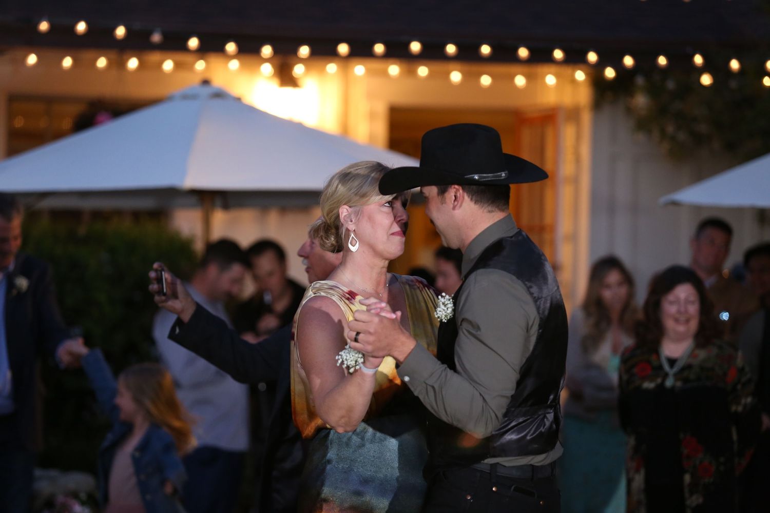 A couple dances together at an evening wedding reception with twinkling lights in the background.
