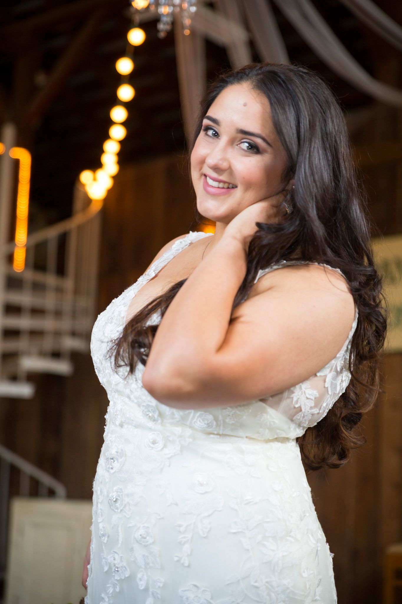 A bride in a white lace dress poses at a wedding venue with decorative string lights in the background.