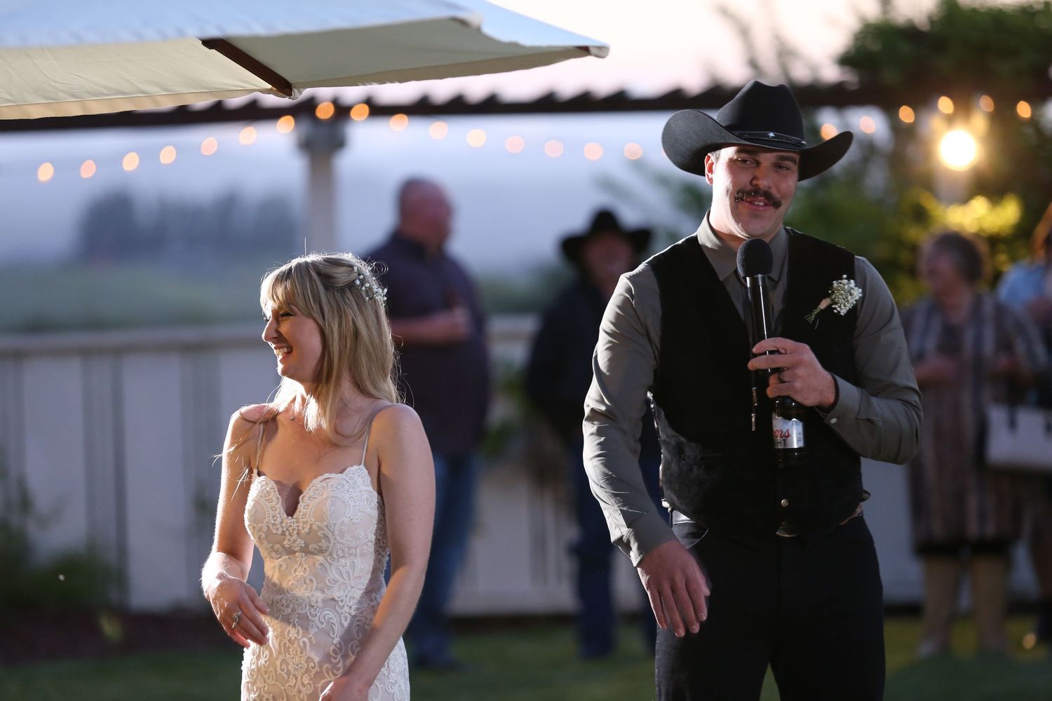 A couple stands outdoors at a wedding ceremony during sunset, with the bride in white and groom in black attire.