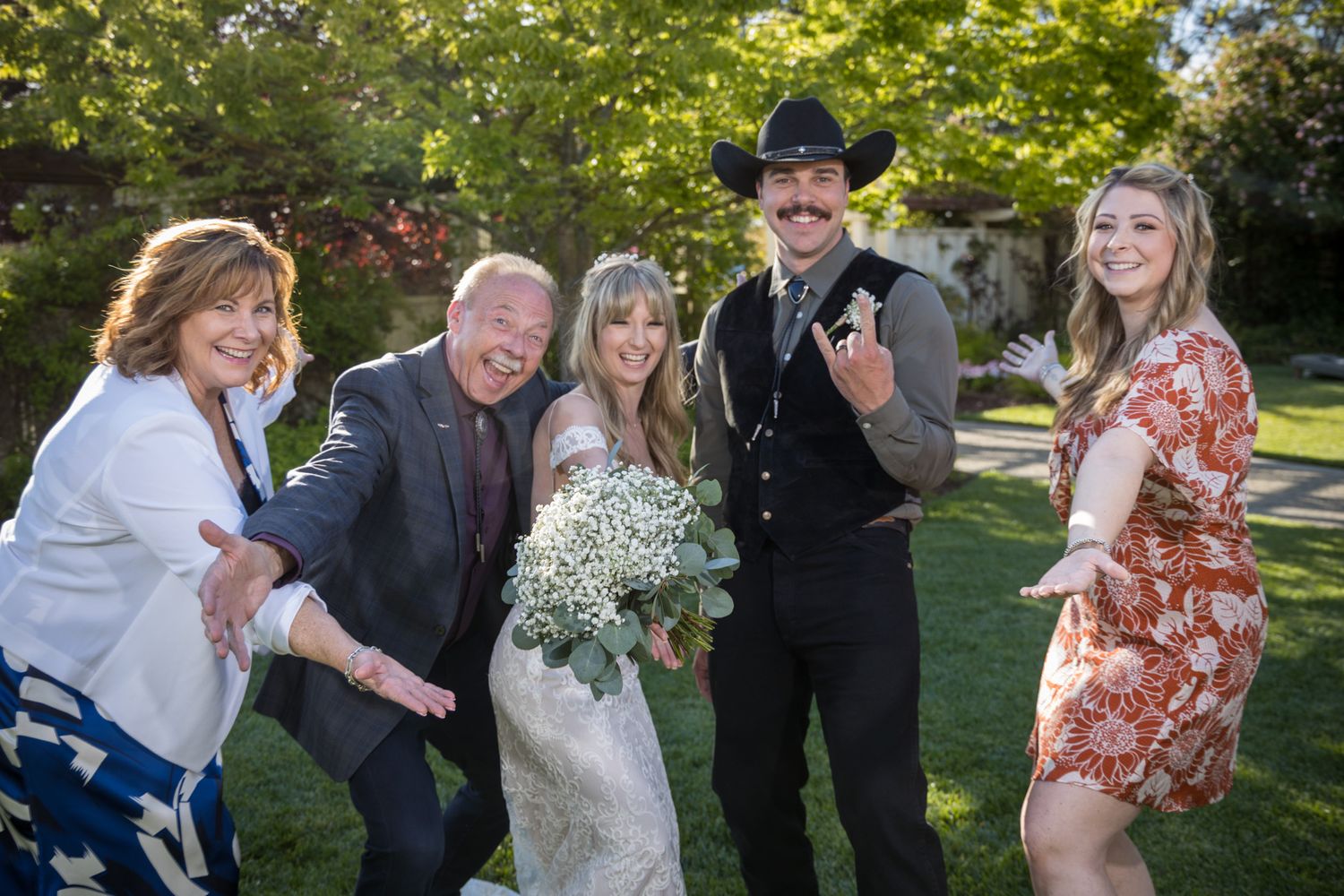 A group of joyful people celebrate outdoors on a sunny day, with a couple dressed in formal attire and floral bouquet.