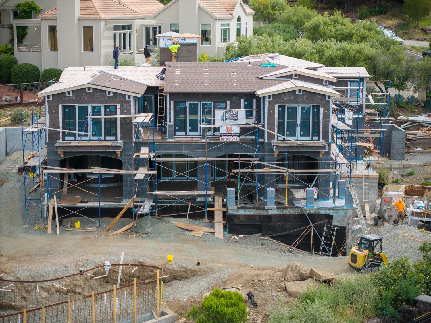 Aerial view of a large house under construction with scaffolding, framing, and building materials on a hillside lot.