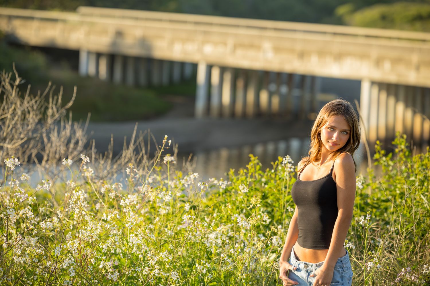 Young woman in black tank top and jeans standing among wildflowers near a bridge in golden hour light.