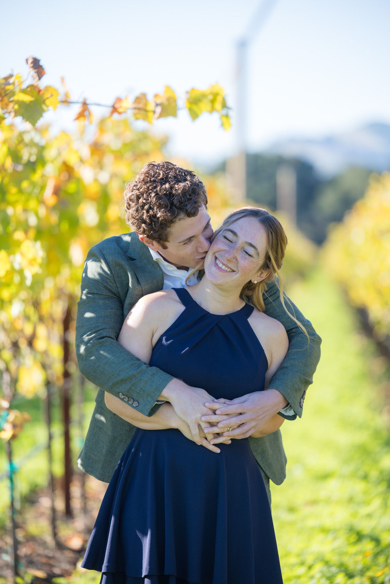 Couple embracing in a vineyard, man kissing woman's cheek as she smiles, surrounded by autumn foliage.