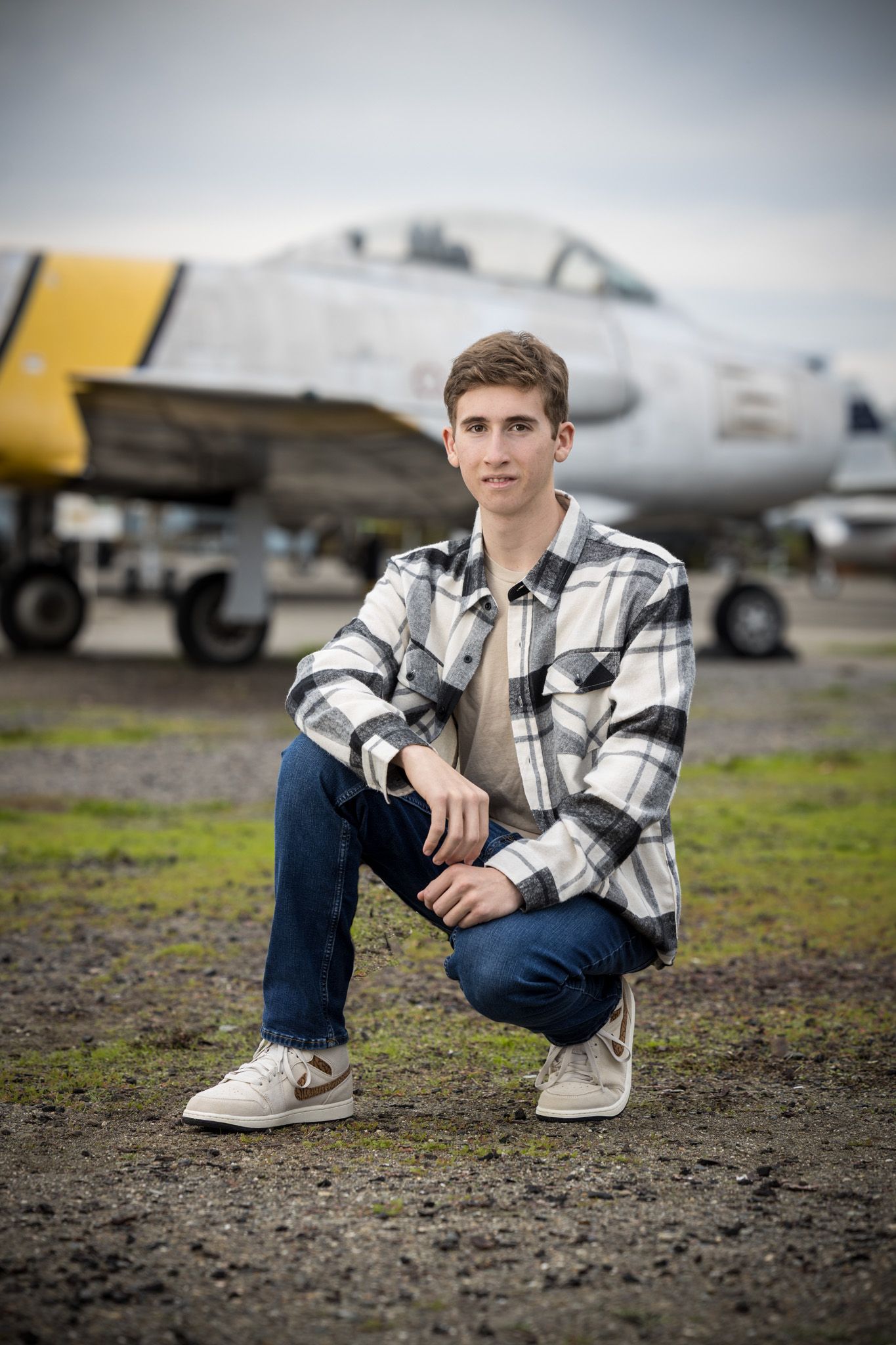 Senior Photos - Kneeling pose next to military aircraft on airfield wearing casual attire.