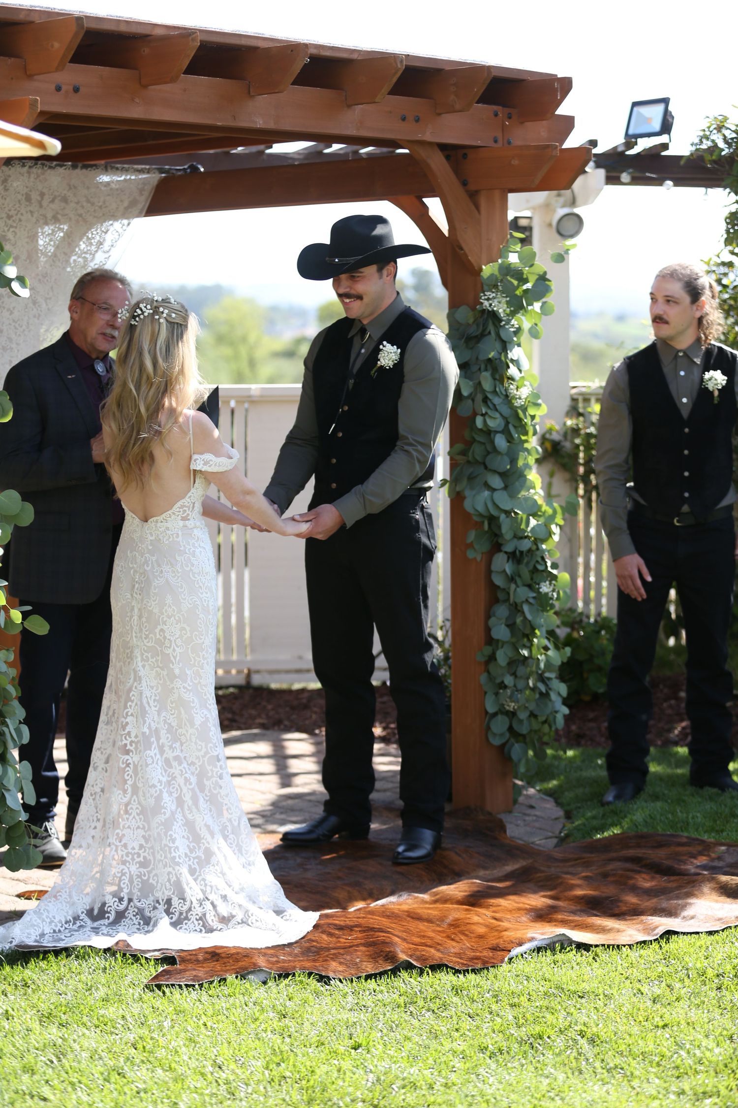 A scenic outdoor wedding ceremony under a wooden pergola decorated with greenery on a sunny day.