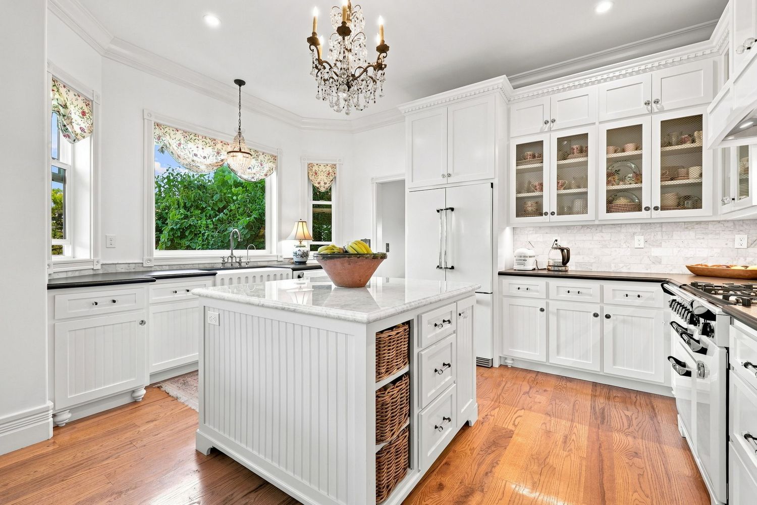 Elegant white kitchen with crystal chandelier, glass-front cabinets, center island with wicker baskets, and hardwood floors.