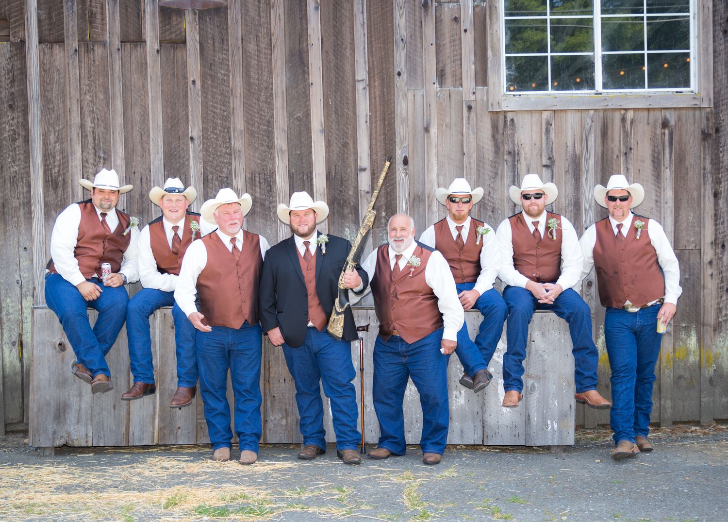 A group in coordinated western attire with cowboy hats, brown vests and jeans poses against a rustic barn wall.