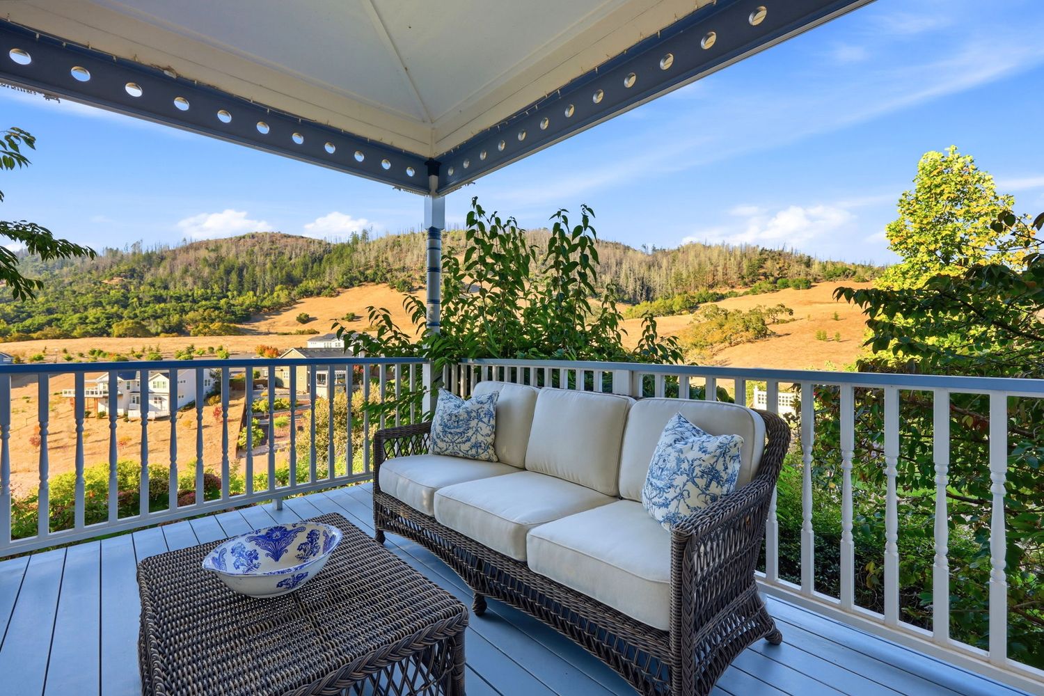 Outdoor deck with wicker sofa and coffee table overlooking scenic rolling hills and lush greenery under a blue sky.