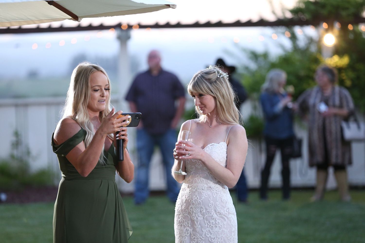 Two women in formal attire stand together at an outdoor event, one wearing a green dress and one in white lace.