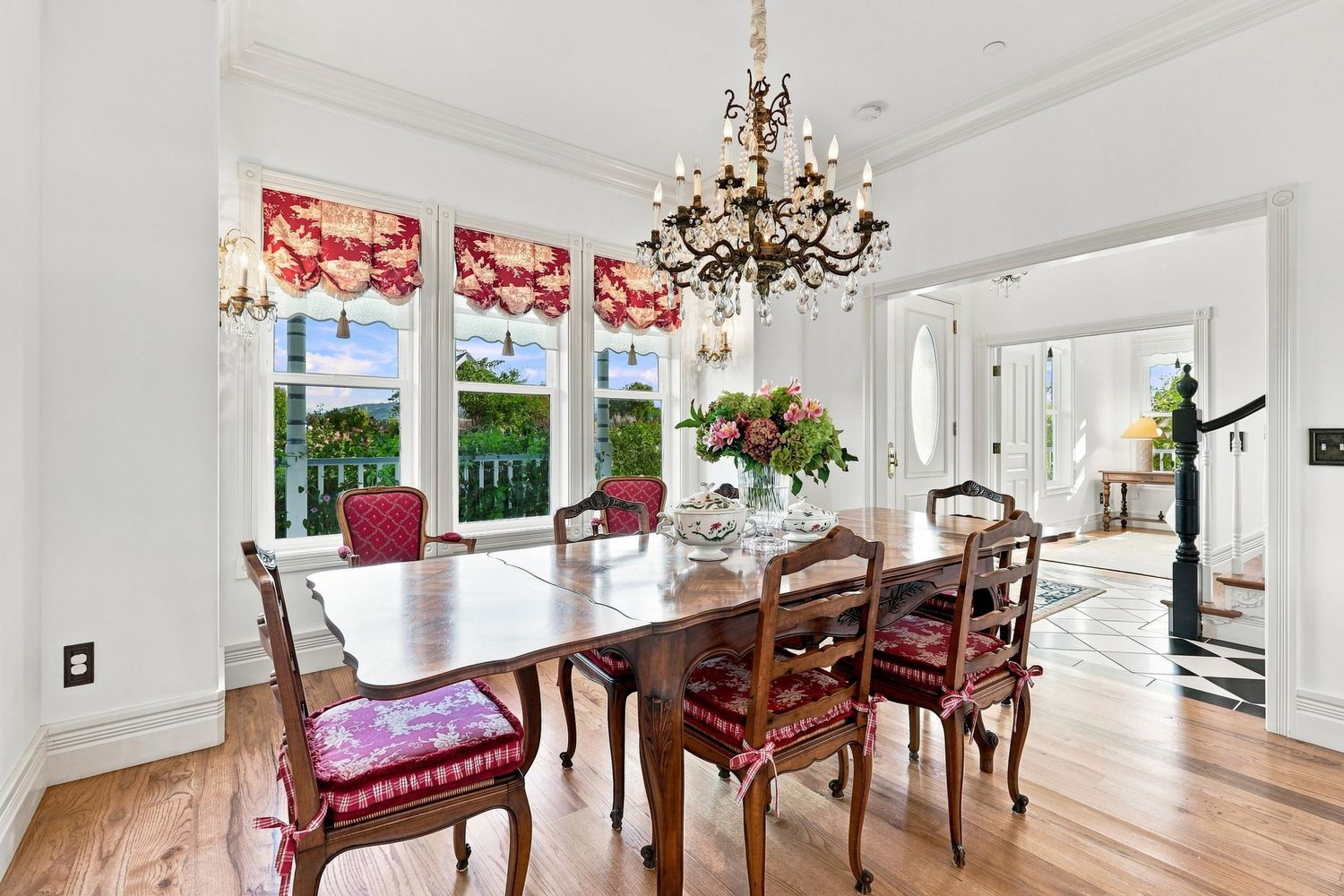 Formal dining room with wood table, red upholstered chairs, chandelier, and large windows with floral valances.