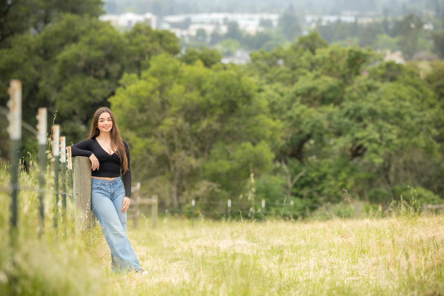 Young woman in jeans and black top poses by a fence in a lush green meadow with trees in the background.