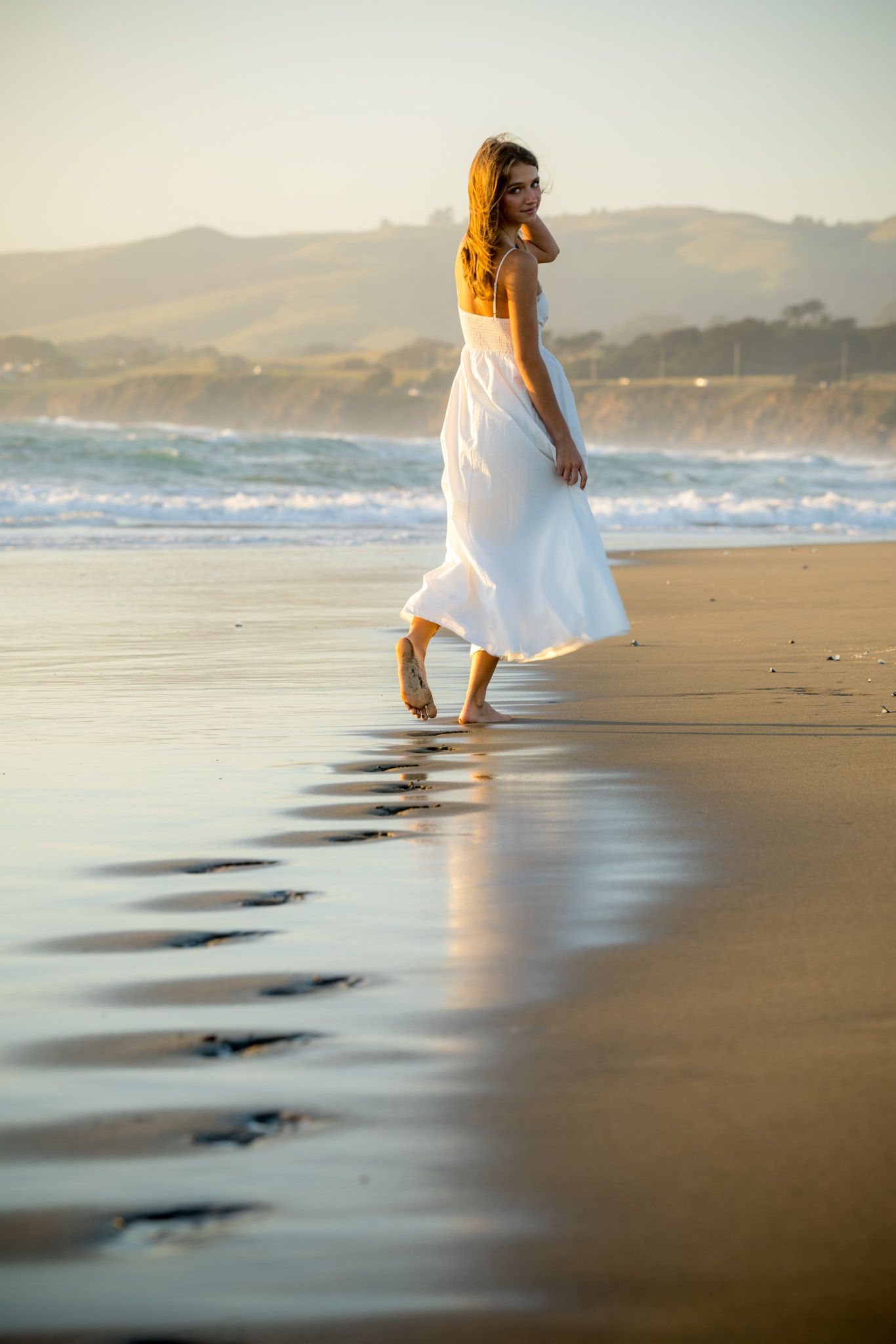 Woman in white dress walking barefoot on a sandy beach at golden hour with ocean waves and mountains in background.