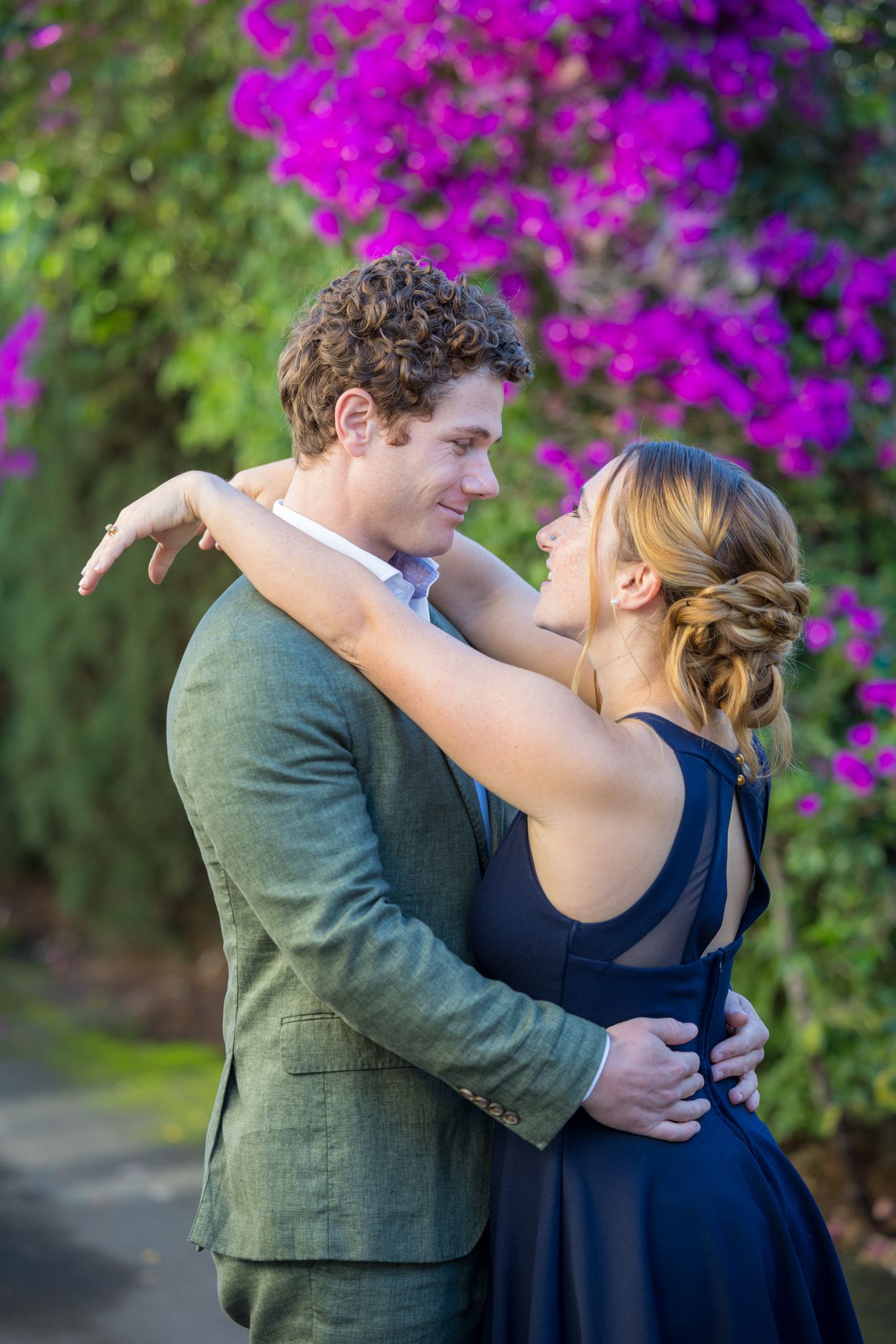 Couple embracing outdoors in formal attire with vibrant pink bougainvillea flowers in the background.