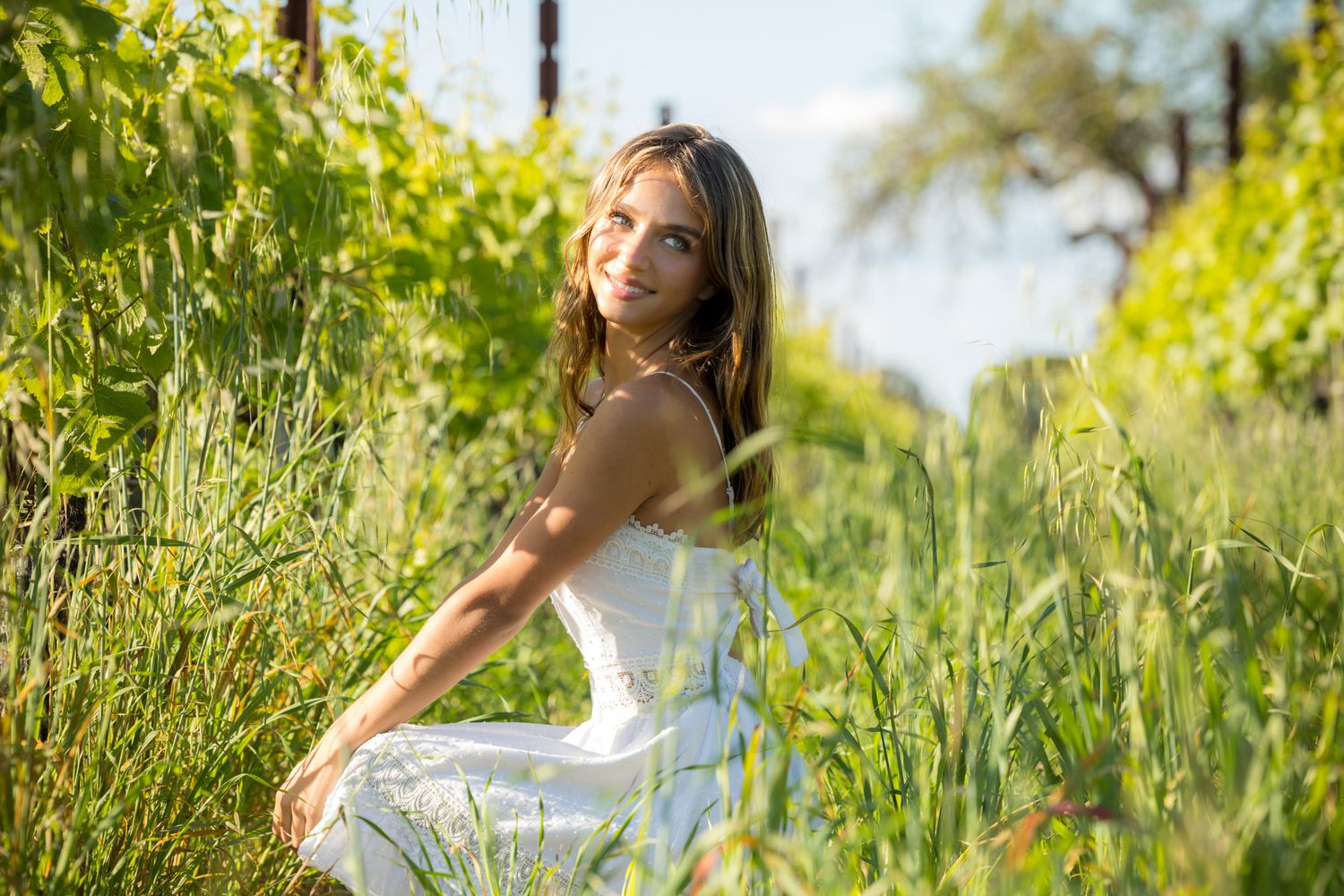 A young woman in a white dress sits smiling among lush green vineyard plants on a sunny day.