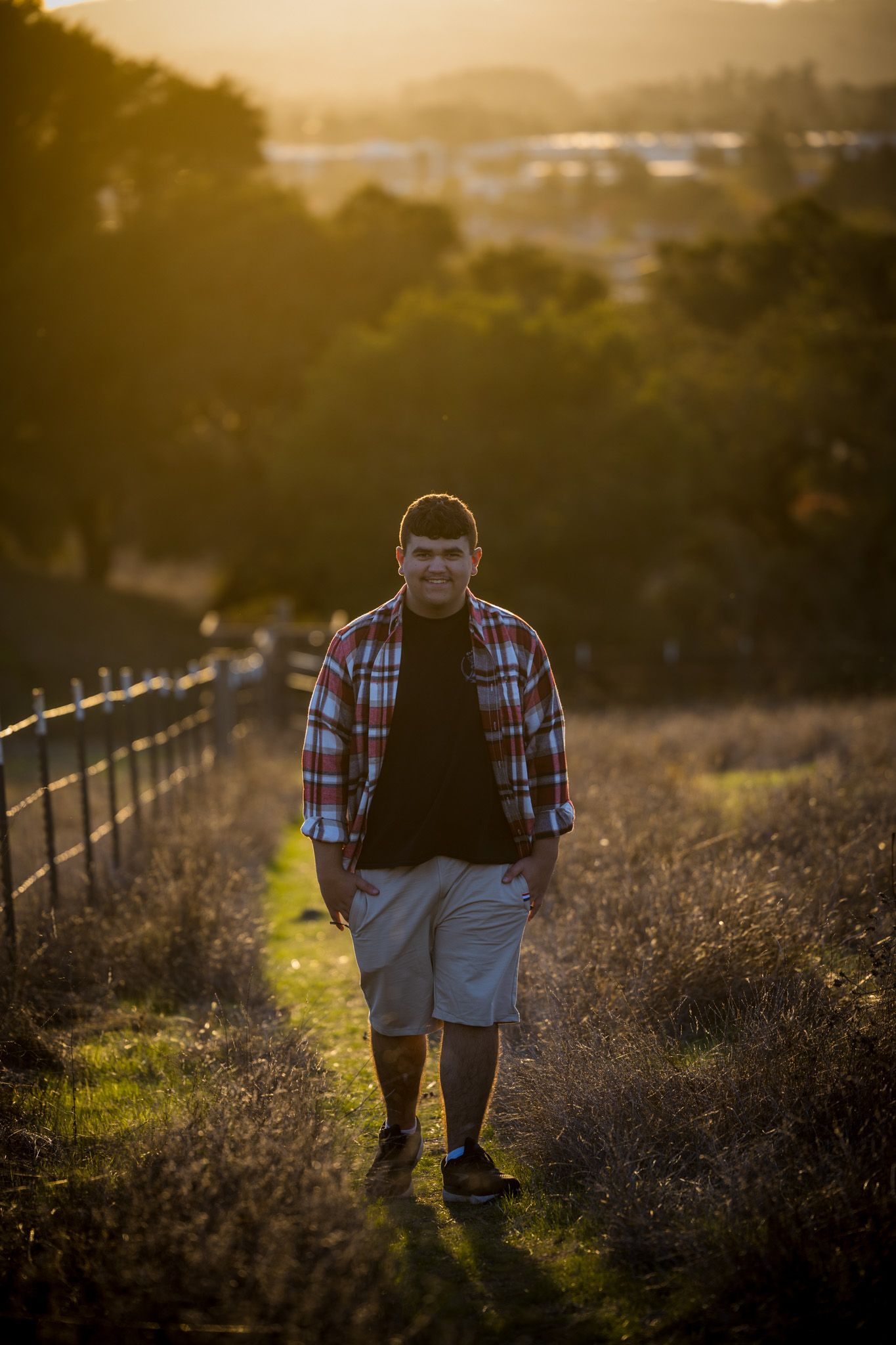 Senior Photos -Subject walks along scenic trail with fence during warm sunset lighting.