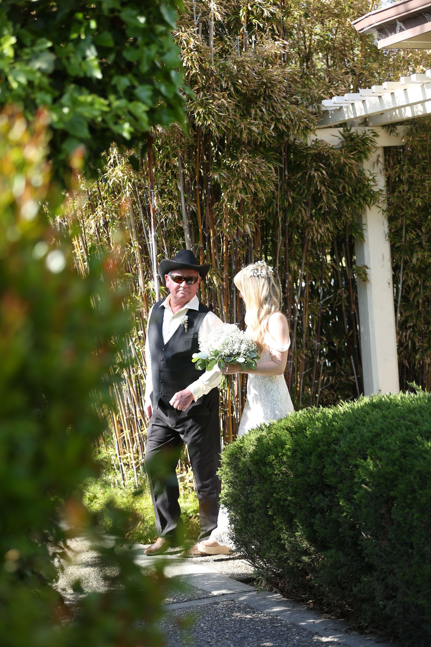 A couple stands together on a garden pathway near bamboo and a white column during a wedding ceremony.