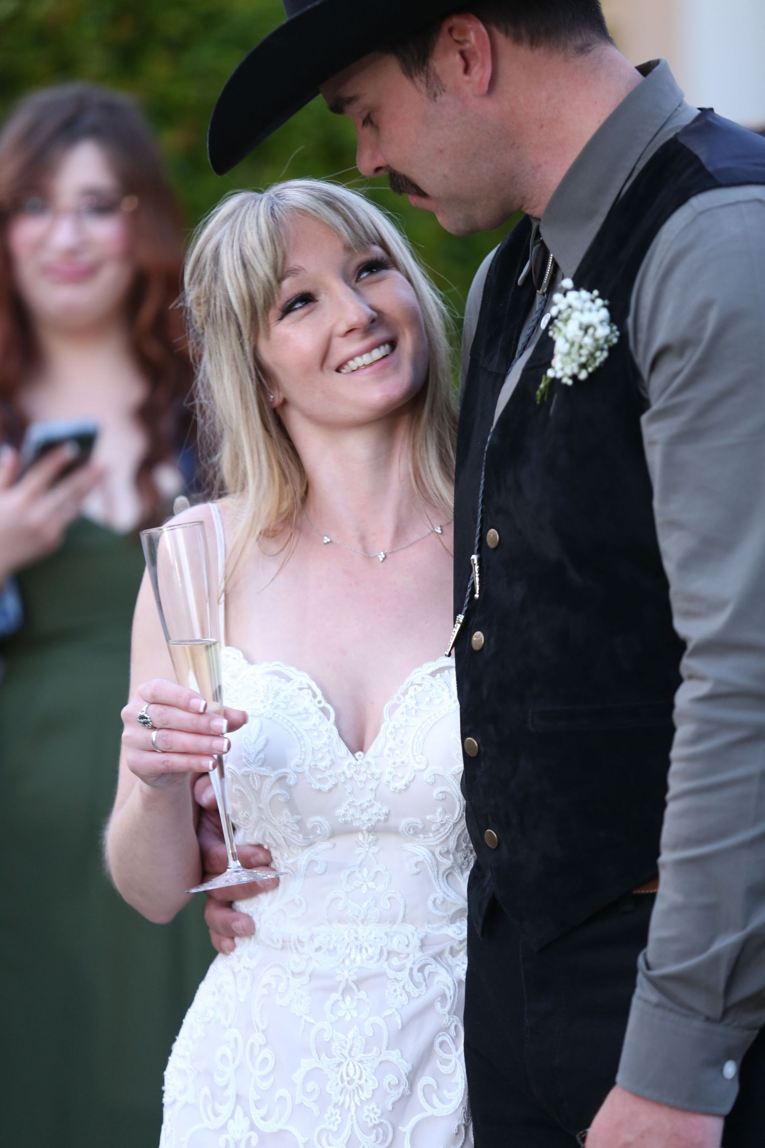 Bride in white lace gown shares tender moments with groom during outdoor wedding reception.