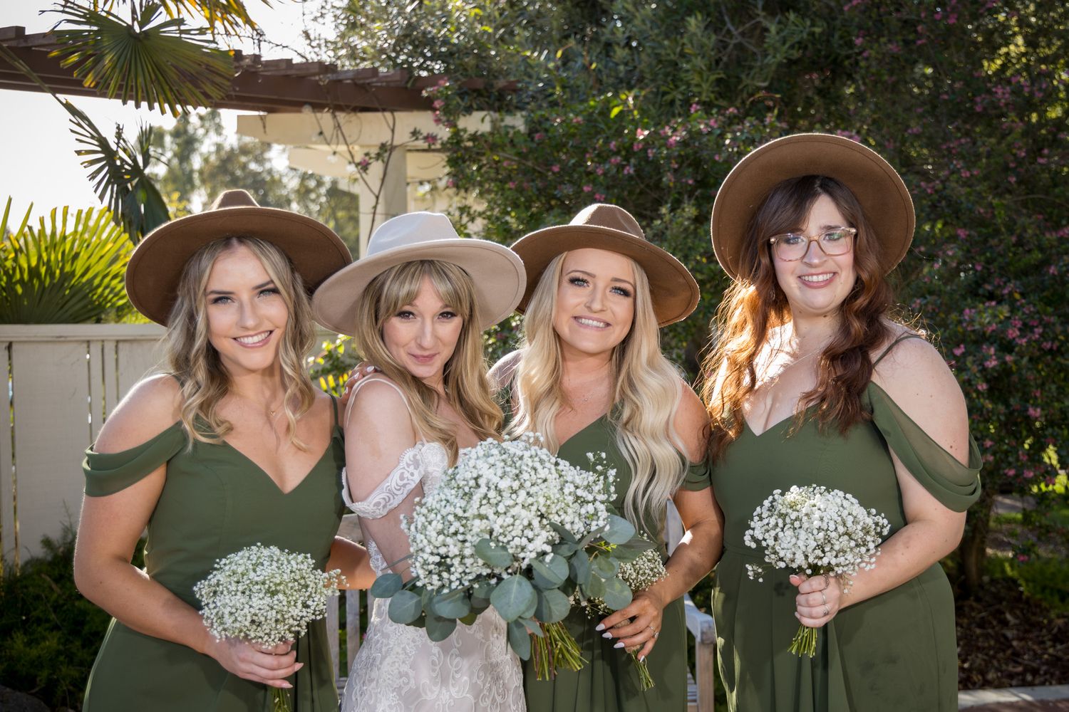 Wedding party wearing sage green dresses and straw hats pose together holding white baby's breath bouquets outdoors.