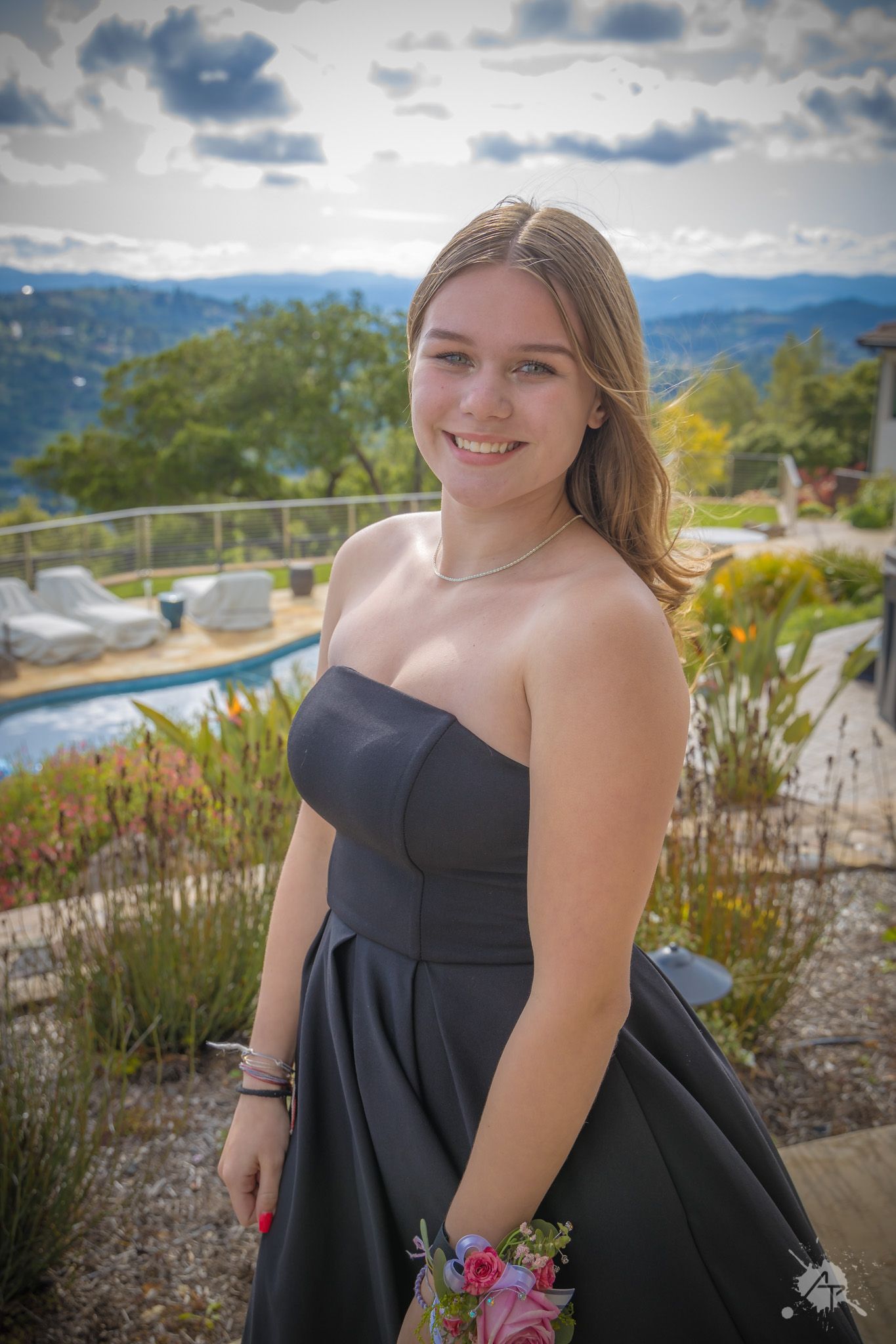 Person in black formal dress holds flowers while posing on scenic mountain overlook.