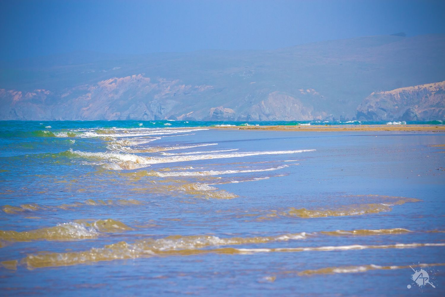 Turquoise ocean waves roll towards a mountainous coastline under bright blue skies on a sunny day.