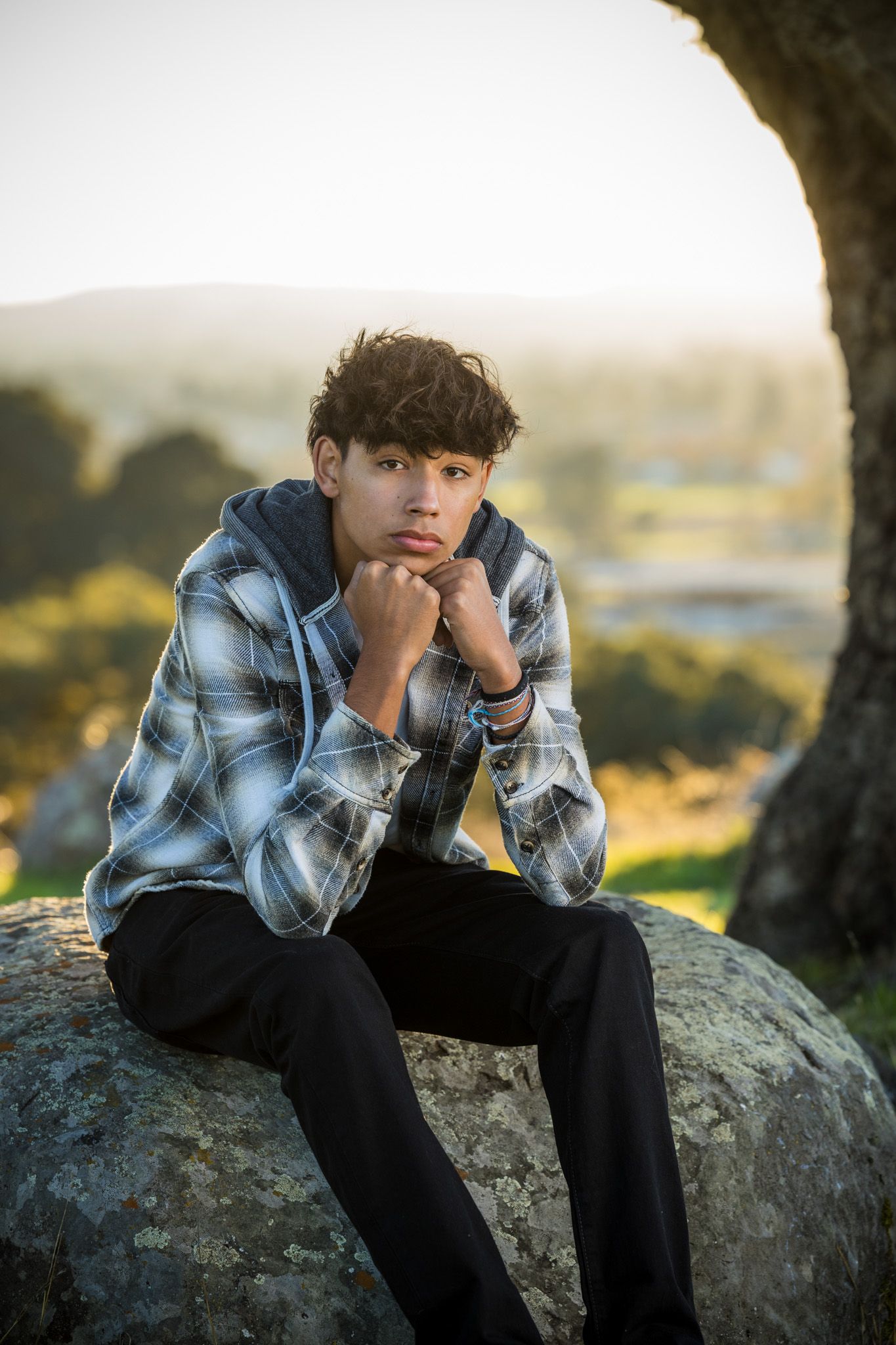 Senior Photos - Moody outdoor portrait of someone sitting on rocks wearing a plaid shirt and black pants.