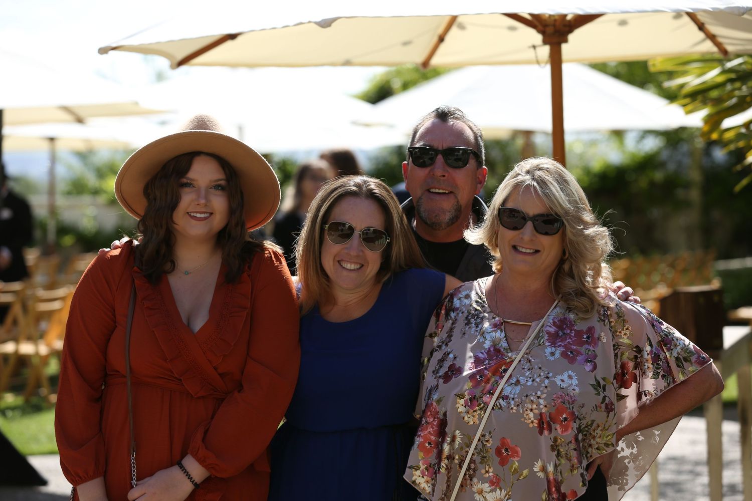 A group stands together smiling outdoors on a sunny day with umbrellas and palm trees visible in the background.