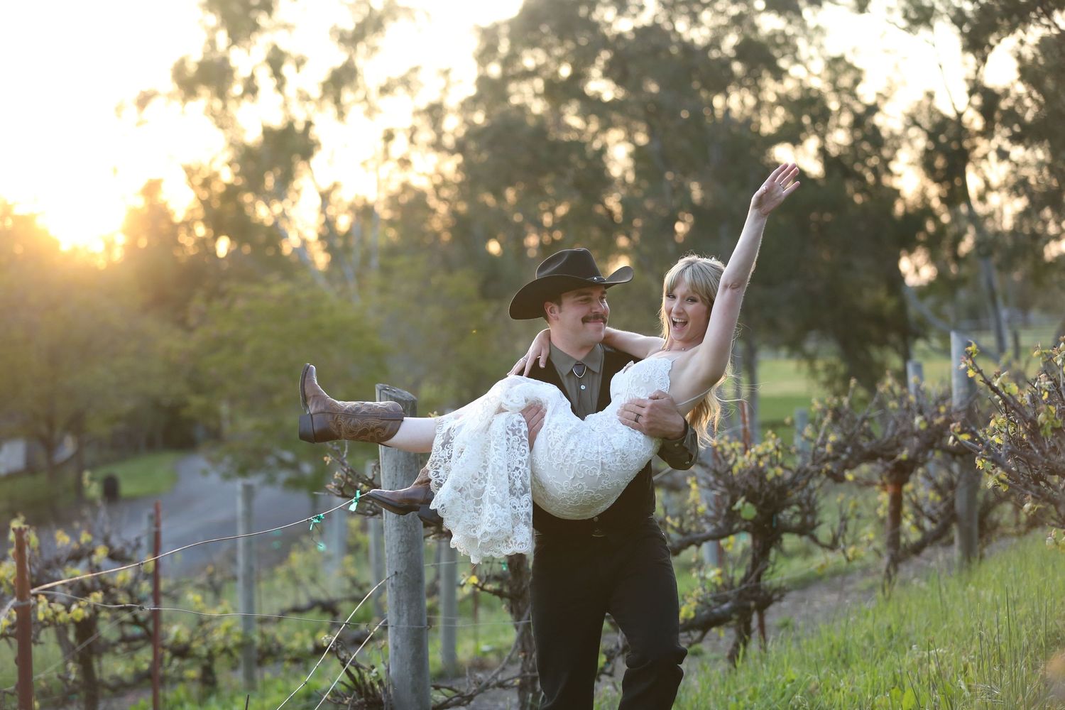 A joyful couple dances together in a sunlit vineyard during golden hour among grape vines on a hillside.