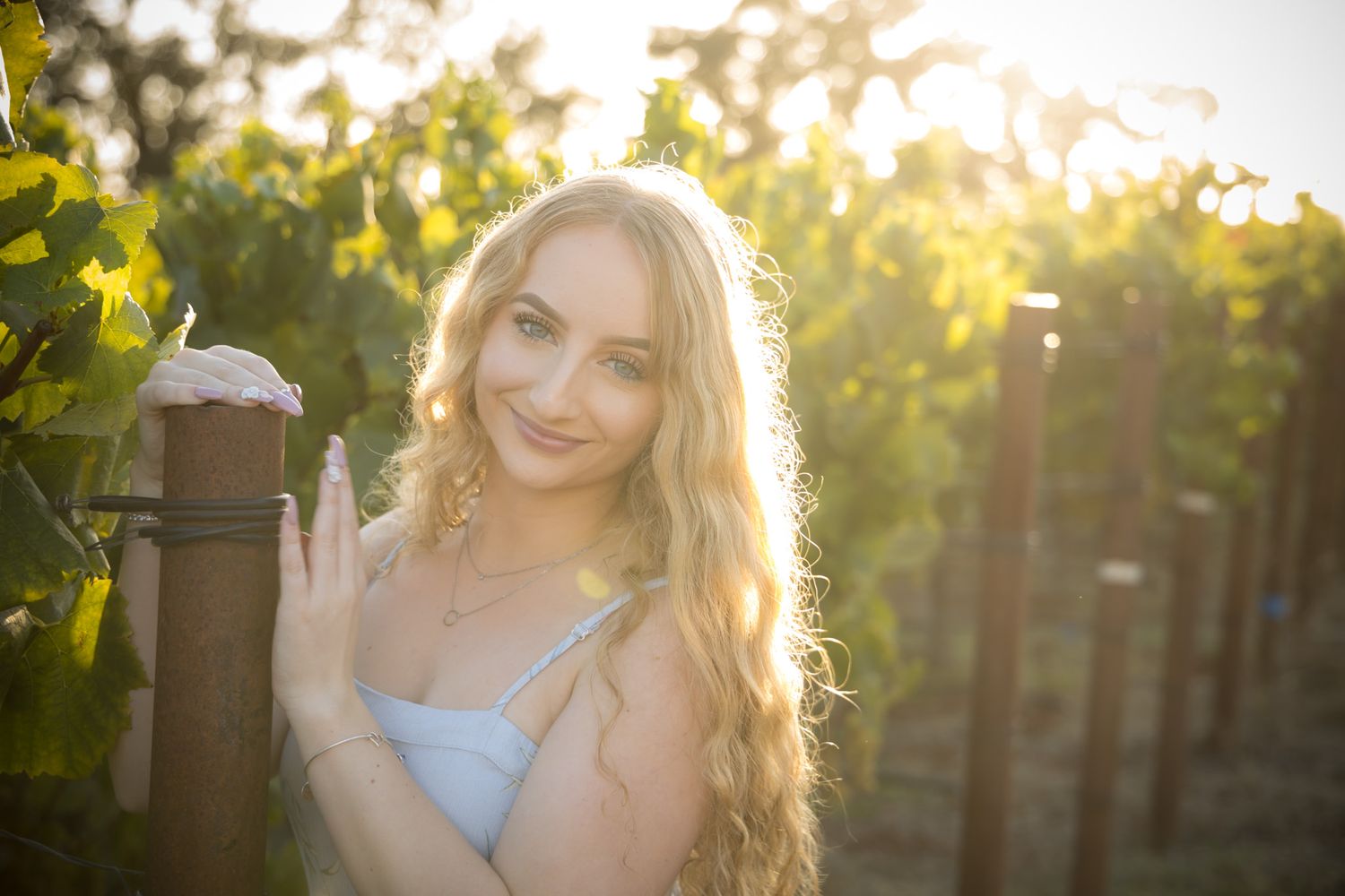 A backlit figure in a white top poses against a wooden post in a vineyard at golden hour.