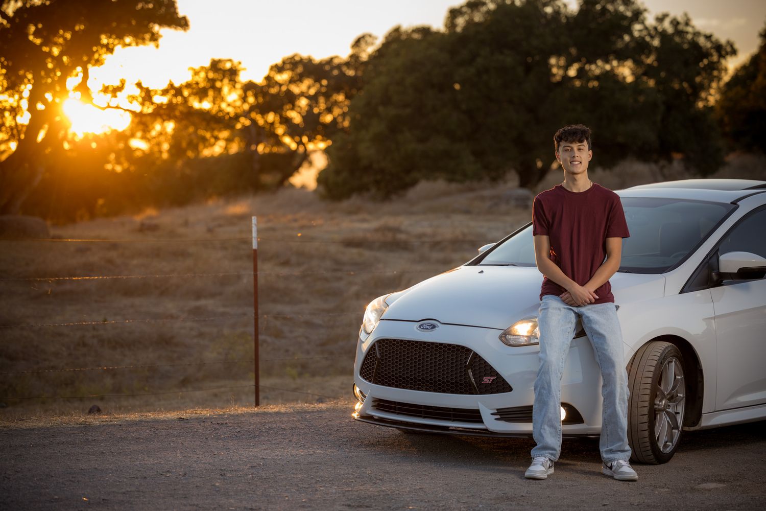 Senior Photos - Person leaning against white car during sunset with rural landscape in background.