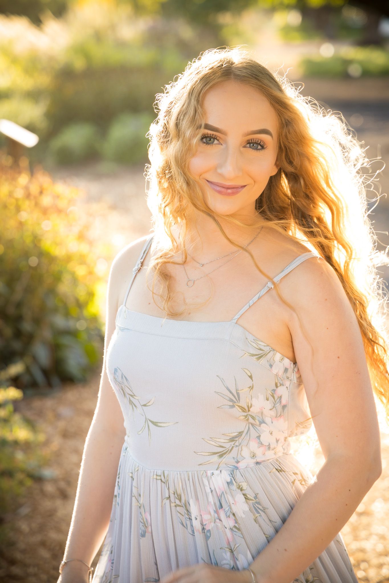 A person in a delicate floral dress poses outdoors during golden hour with sunlight creating a warm backlit glow.