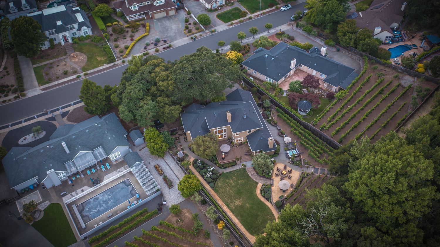 Aerial drone view of upscale residential properties with manicured gardens, slate roofs, and tree-lined streets in a suburban neighborhood.