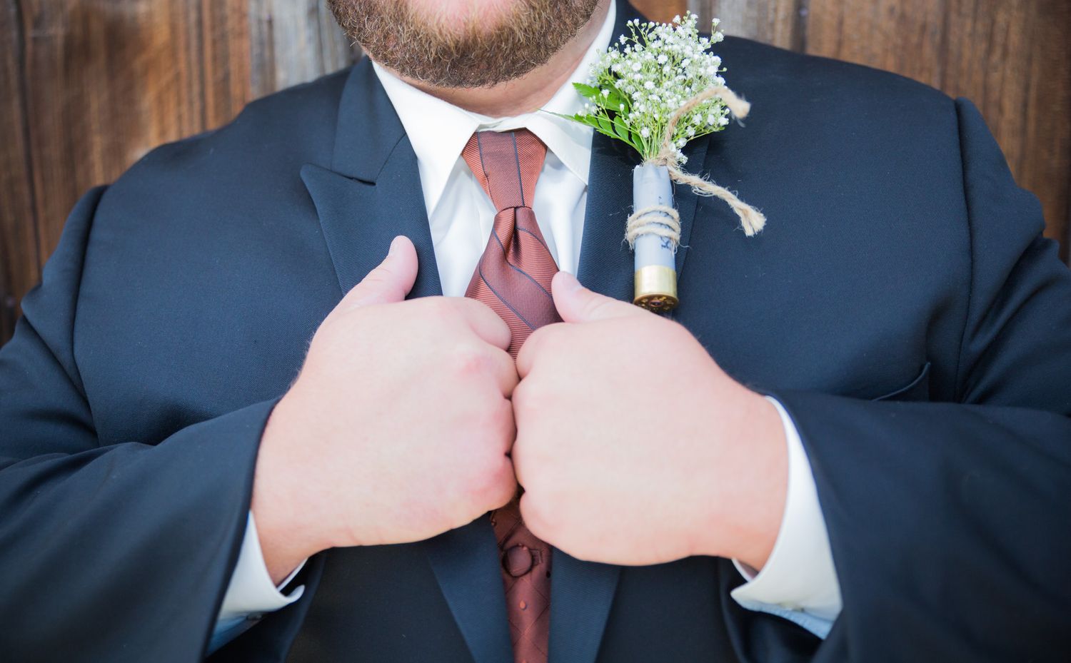 Close-up view of a groom in navy suit and burgundy tie adjusting his outfit with a small white boutonniere.