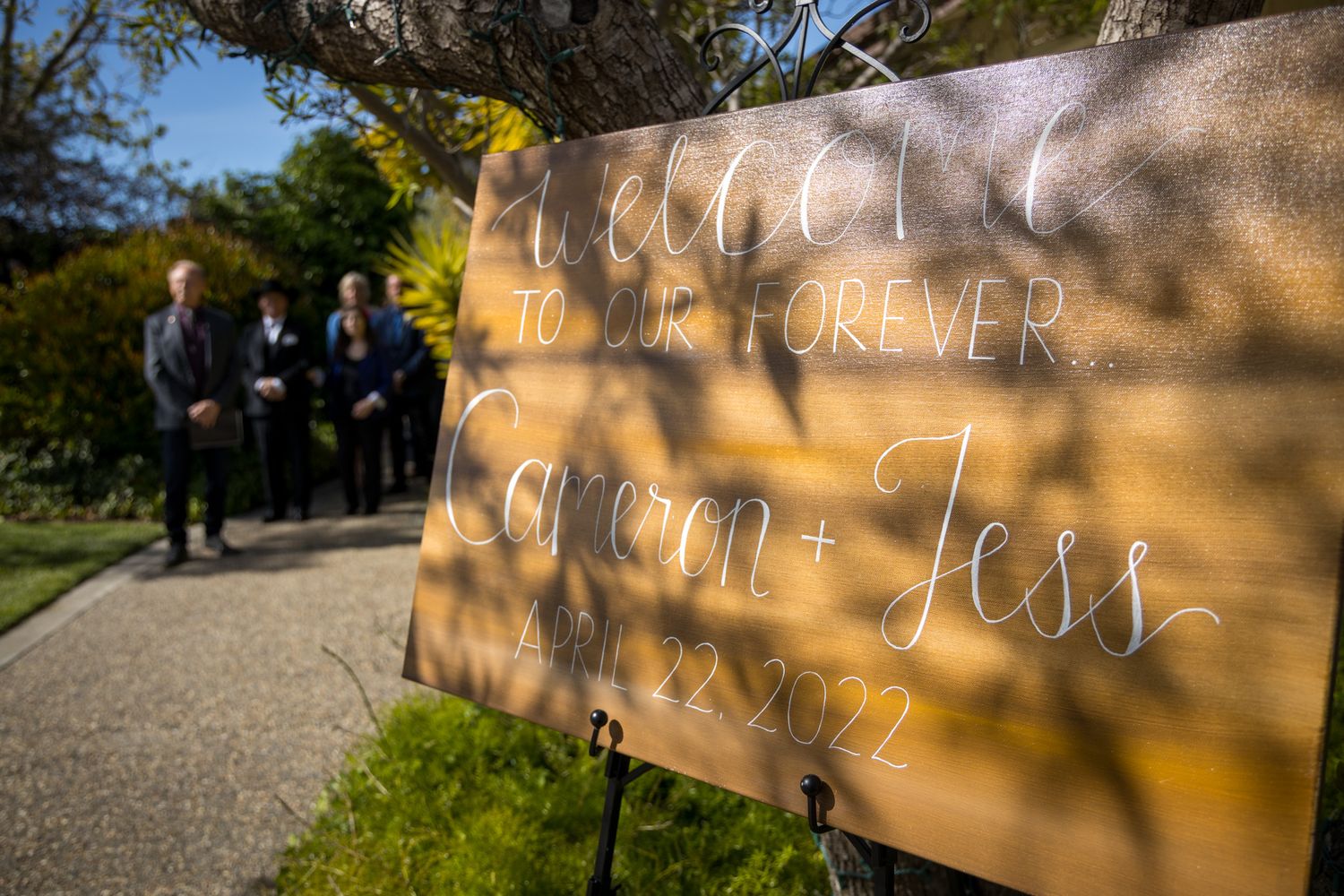 A rustic wooden wedding sign with text photographed outdoors with blurred guests in the background.