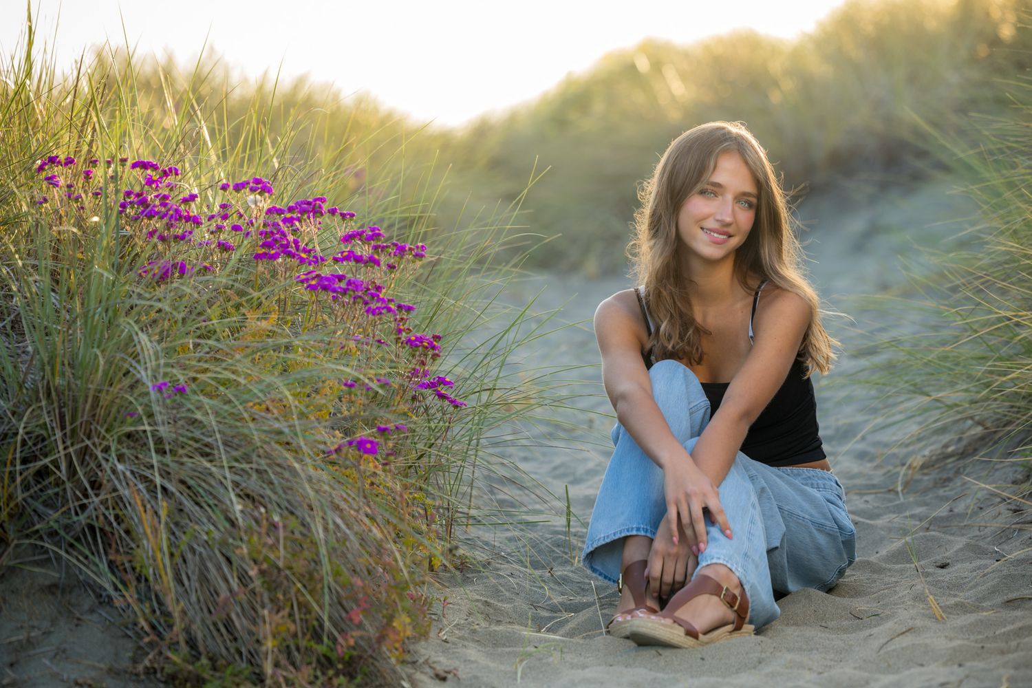 Young woman crouching on sandy dunes beside purple wildflowers and tall grass in warm golden sunlight.