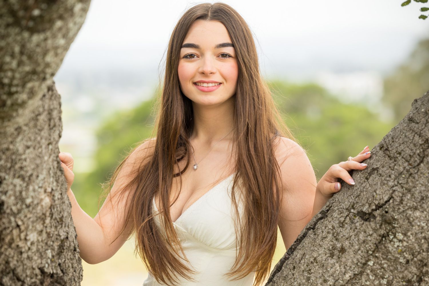 Young woman with long brown hair smiling outdoors, leaning against a tree with a blurred green landscape background.