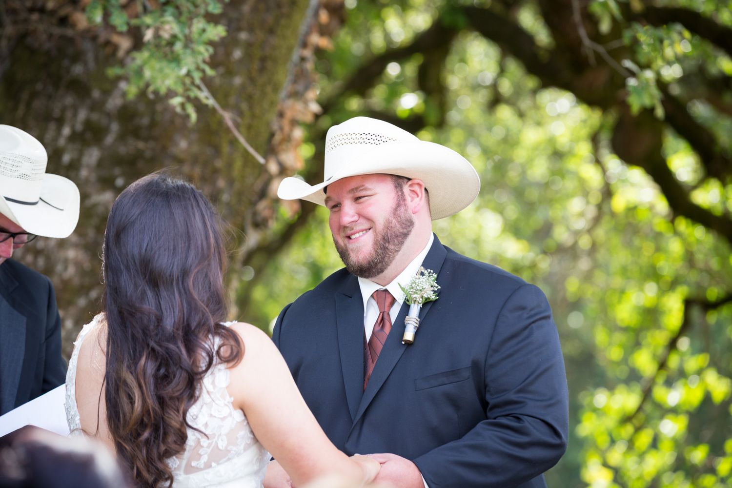 Two people share a joyful moment at an outdoor wedding ceremony under a canopy of green trees.