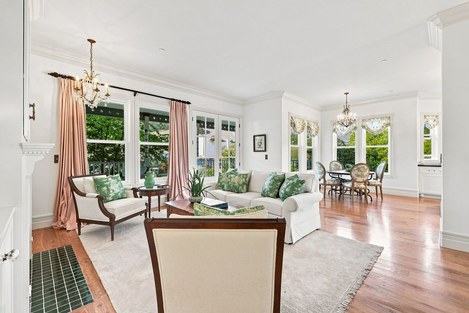 Elegant living room with hardwood floors, pink curtains, green accents, chandelier, and French doors leading to a sunlit dining area.
