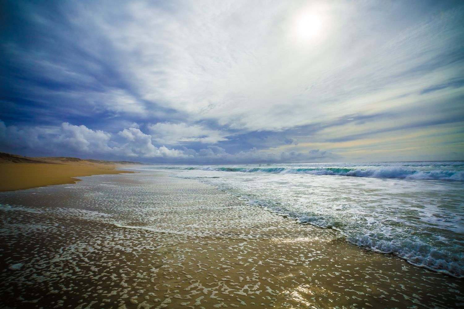Dramatic seascape with rolling waves meeting sandy shore under wispy clouds and bright sunlight.