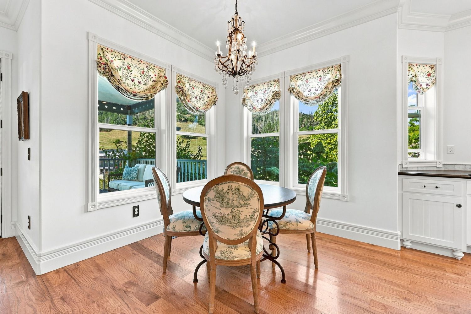 Bright dining room with hardwood floors, chandelier, large windows with floral valances, and a round glass table with ornate chairs.