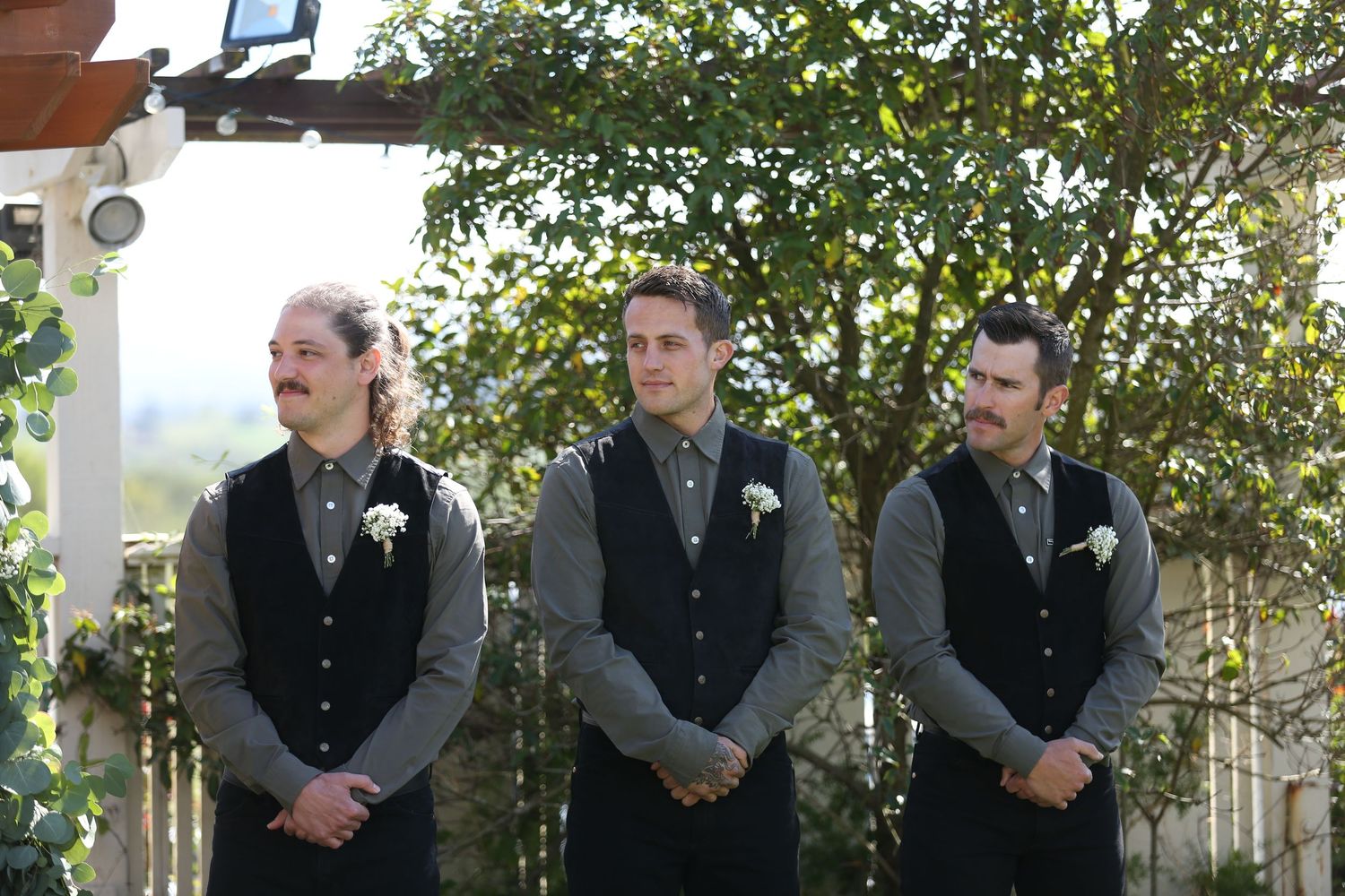 Groomsmen in black suits and gray scarves stand together for wedding party photos by trees and greenery.