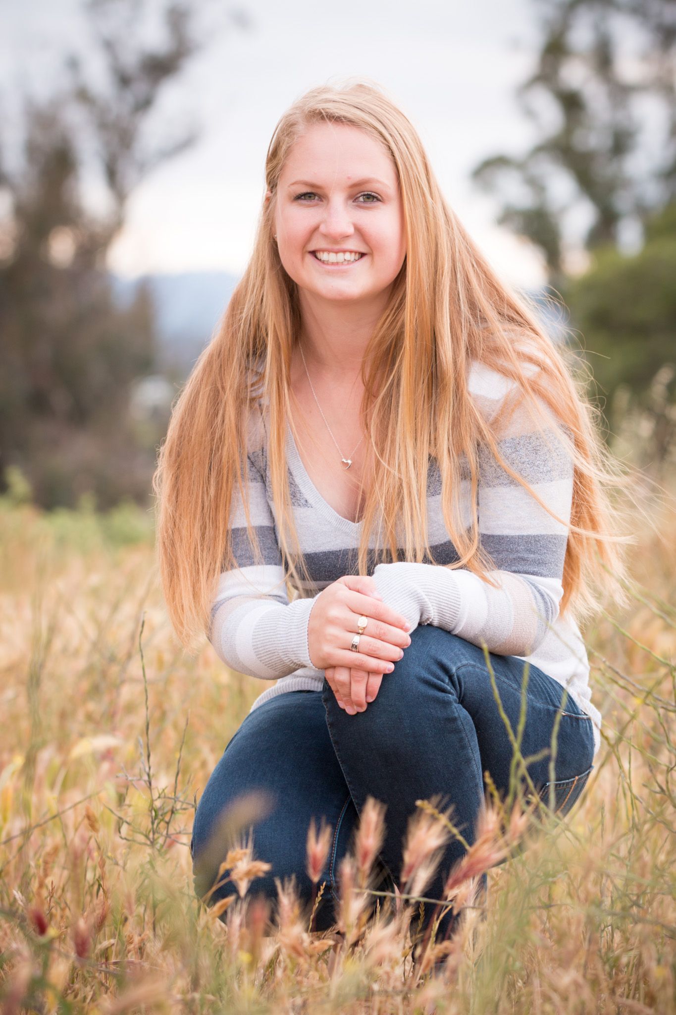 Person crouching in a sunny meadow wearing a gray striped sweater and jeans with long blonde hair.